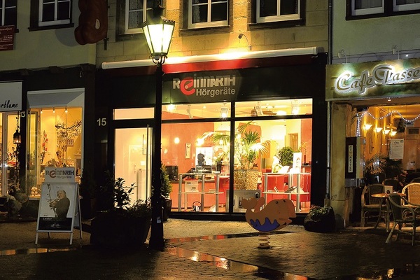 Abendliche Straßenansicht mit beleuchtetem Hörgeräte-Fachgeschäft und angrenzendem Café.