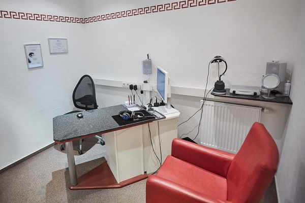 Modern eingerichtetes Hörakustiker-Beratungszimmer mit Schreibtisch, PC und rotem Sessel.